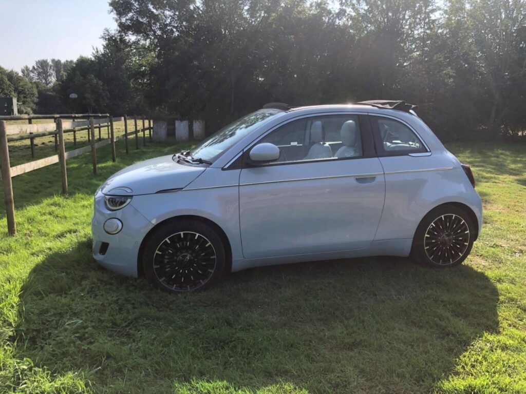 Side profile view of a celestial blue FIAT 500e Cabriolet parked on grass next to a wooden fence