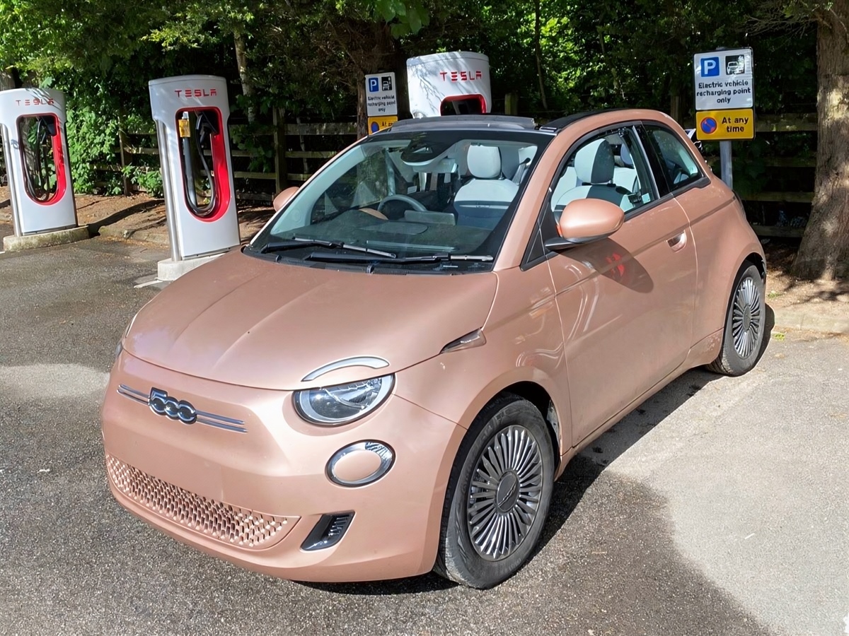 Front three-quarter view of a Rose Gold FIAT 500e La Prima parked at a Tesla Supercharger site.