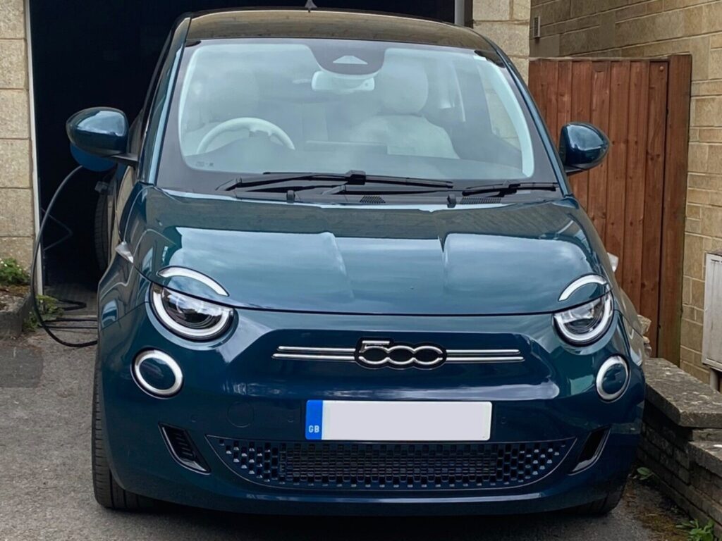 Front view of an ocean green FIAT 500e parked on a driveway with a charging cable connected