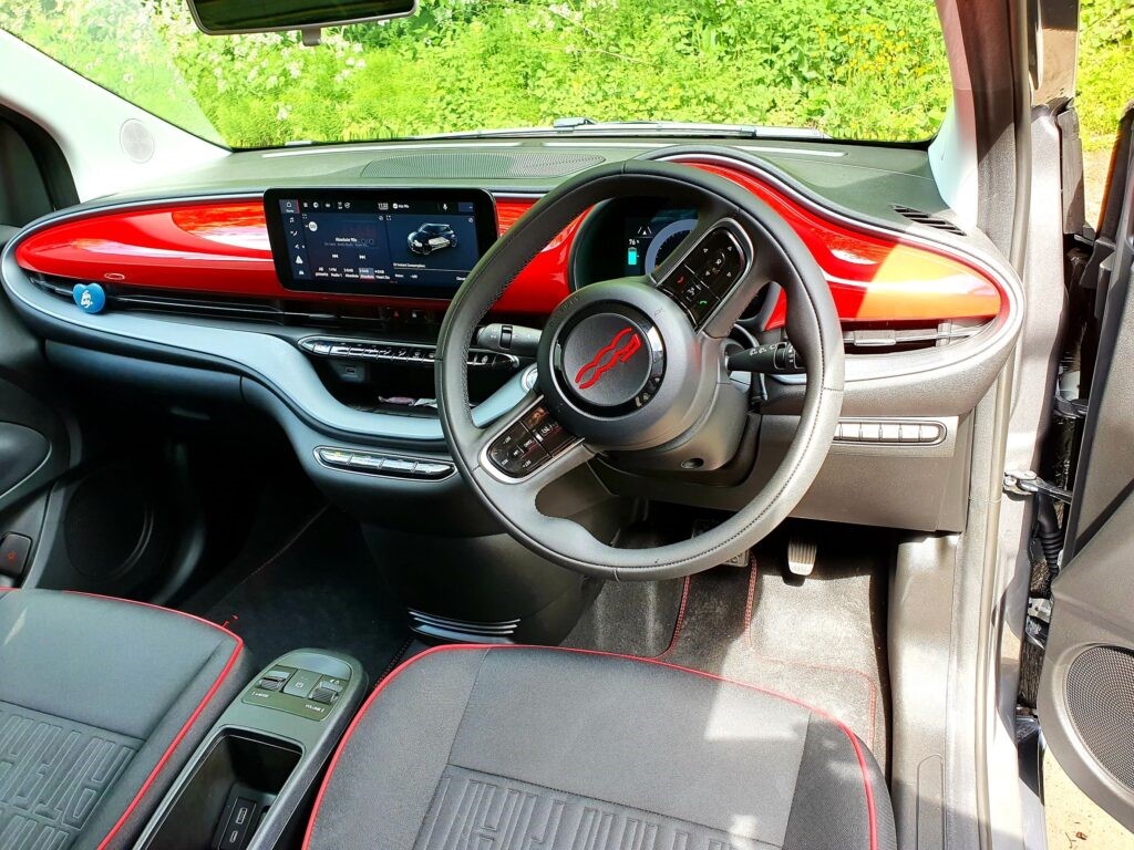 Right-hand drive interior of a FIAT 500e showing the steering wheel, red dashboard trim, and central touchscreen display